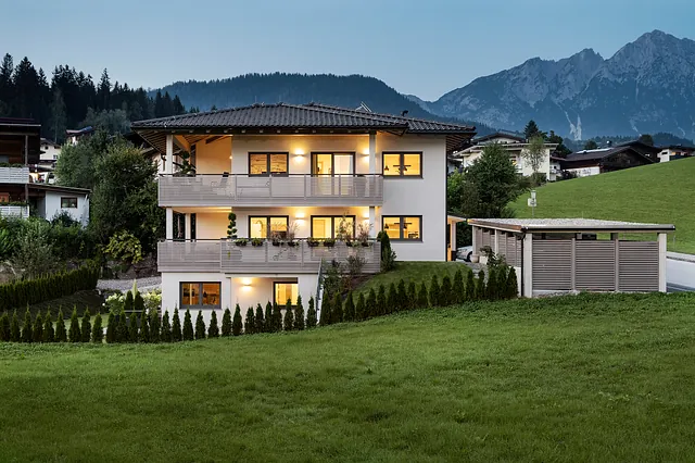 Modernes Apartmenthaus mit drei Etagen, großer Gartenanlage und überdachtem Carport, in Hanglage mit Blick auf den Wilden Kaiser in der Abenddämmerung.
